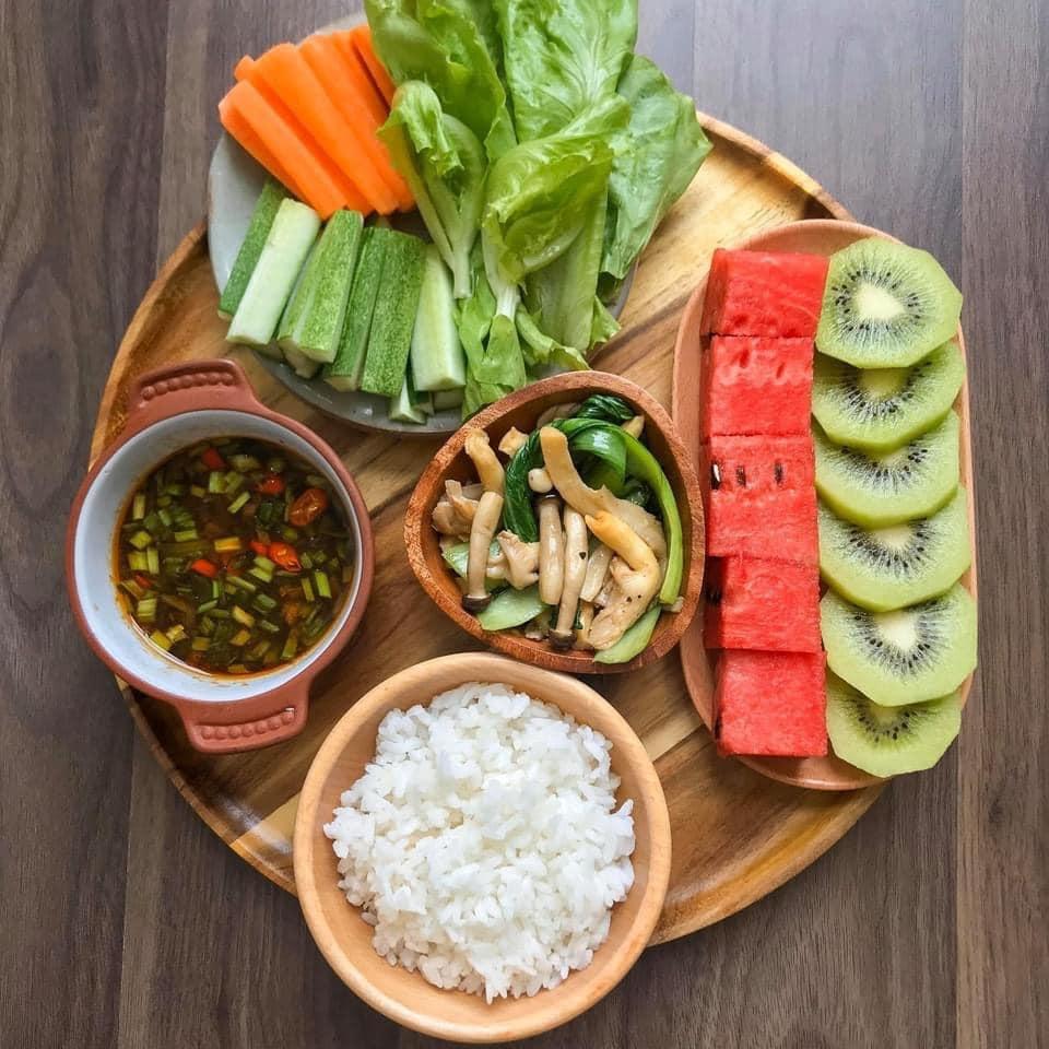 A wooden tray with fresh carrot and cucumber sticks, lettuce, mushrooms, white rice, dipping sauce, watermelon and kiwi slices